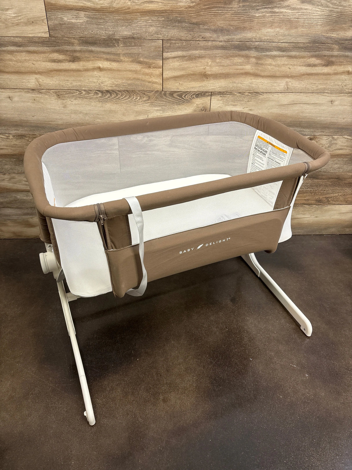 Angled view of Baby Delight Beside Me Dreamer bassinet in mocha showing mesh sides and white metal legs on wood floor