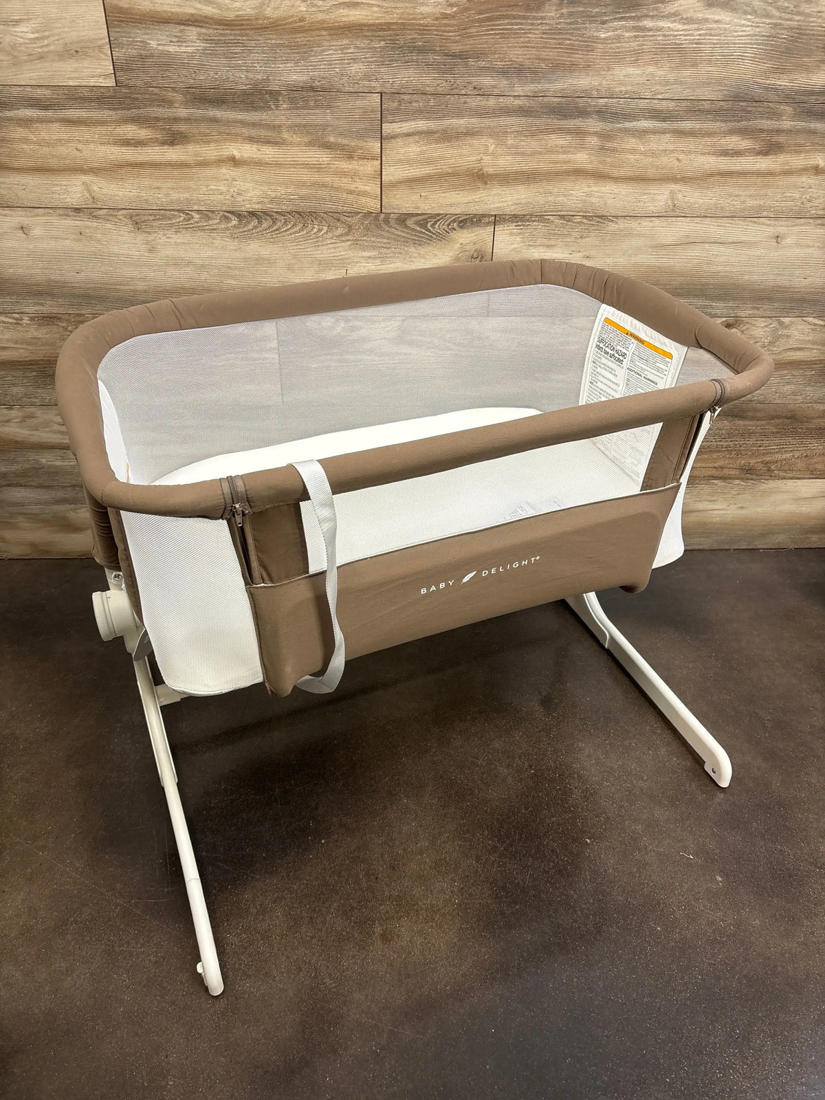 Angled view of Baby Delight Beside Me Dreamer bassinet in mocha showing mesh sides and white metal legs on wood floor
