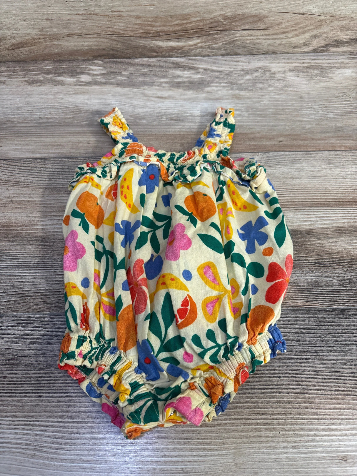 Flat lay of yellow Old Navy smocked bubble romper with ruffle trim and multicolor floral print on wooden background, size 3 to 6 months