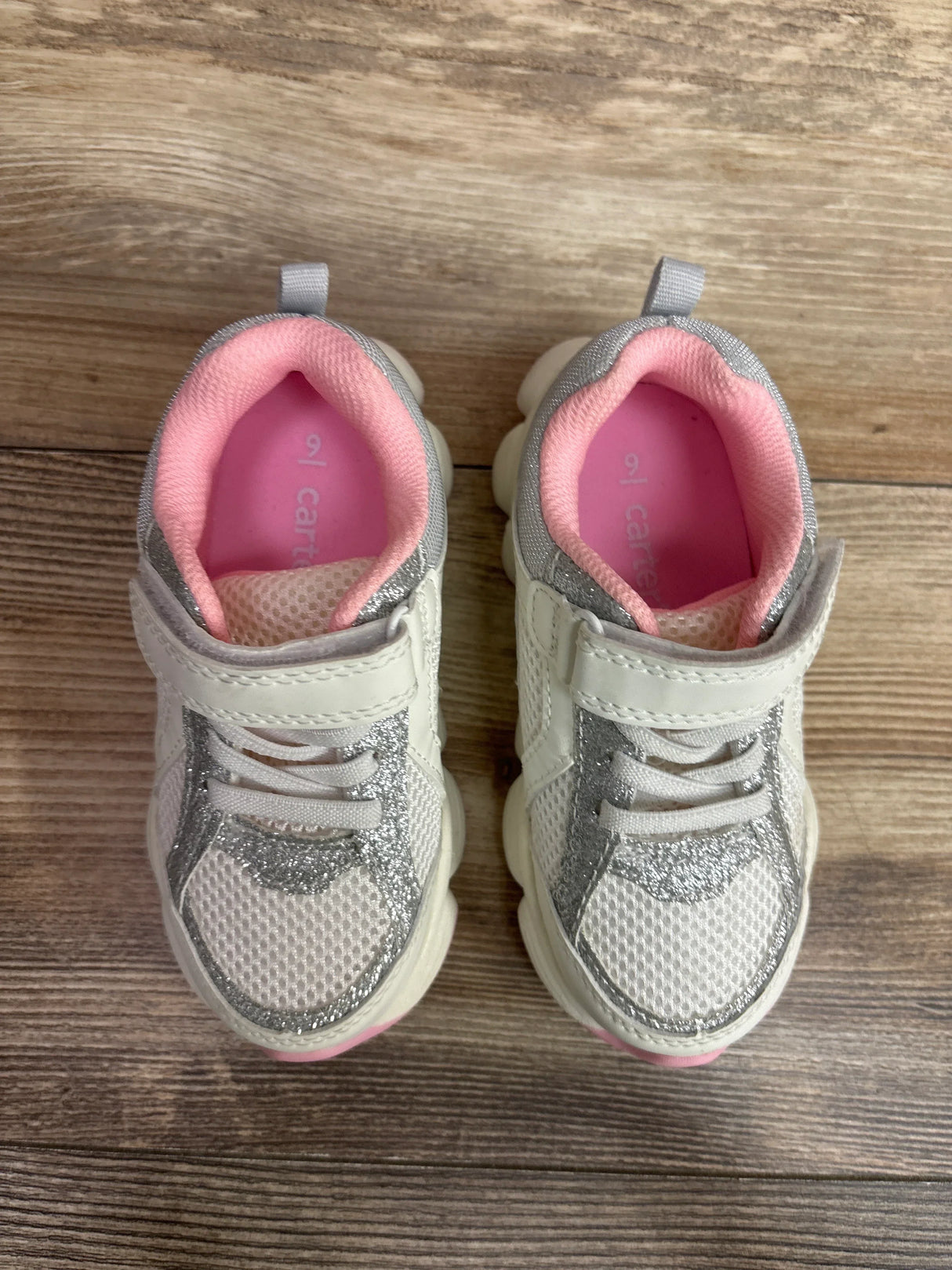 Overhead view of Carter's toddler sneakers in white with silver glitter accents, pink insoles, mesh upper and Velcro on wood floor