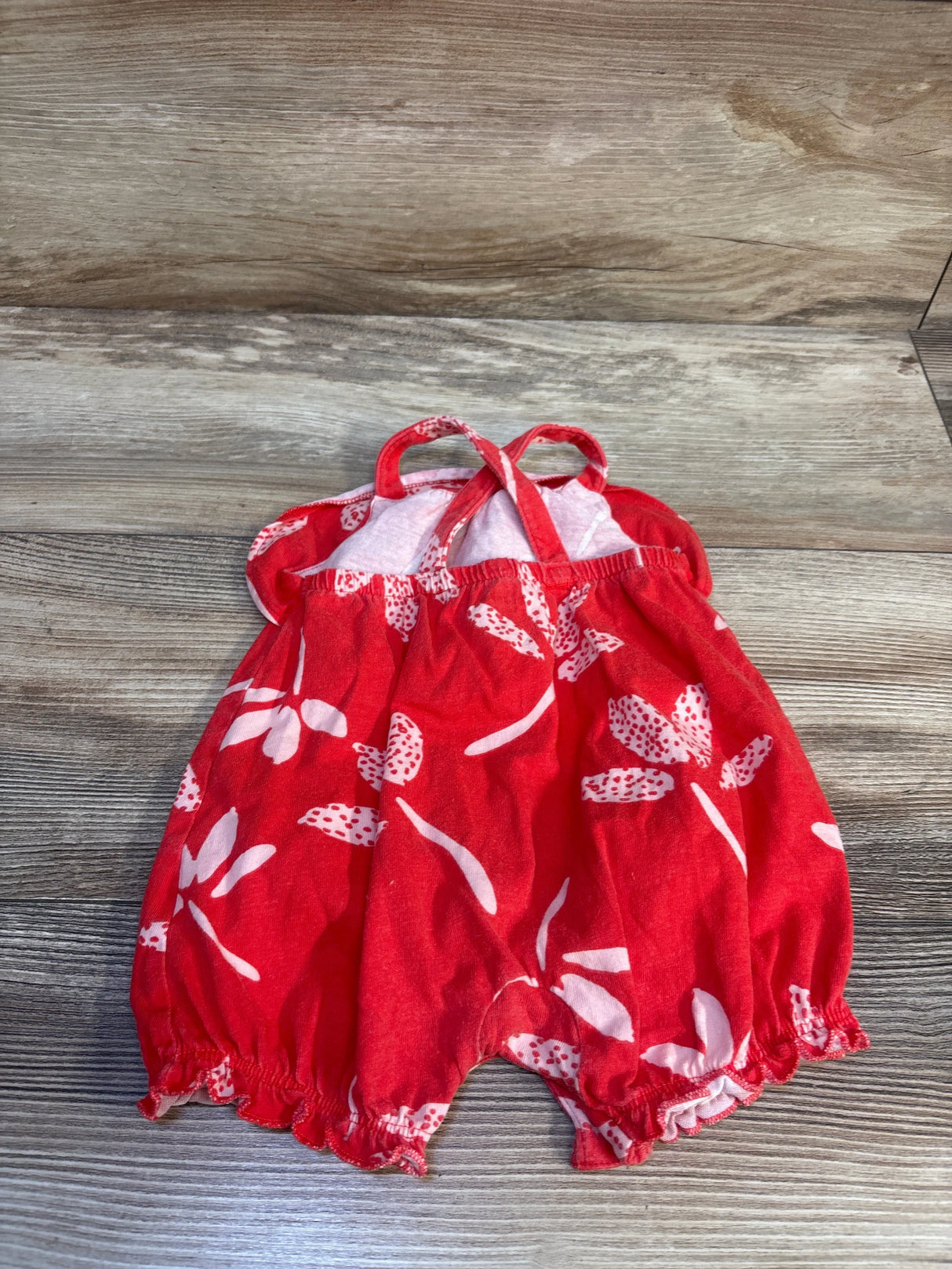 Front view of red floral shortie romper, size 3M, showing elastic gathered leg openings and crossed shoulder straps on wood surface