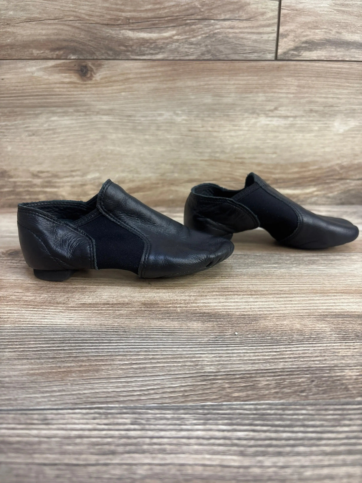 Side view of Capezio EJ2C girls’ black jazz slip-on shoes showing leather upper, elastic panels and low heel on wood