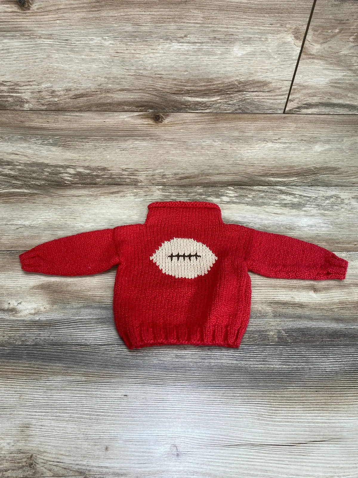 Flat lay back view of red knit baby sweater size 12 months showcasing white football motif with brown stitching on wood floor