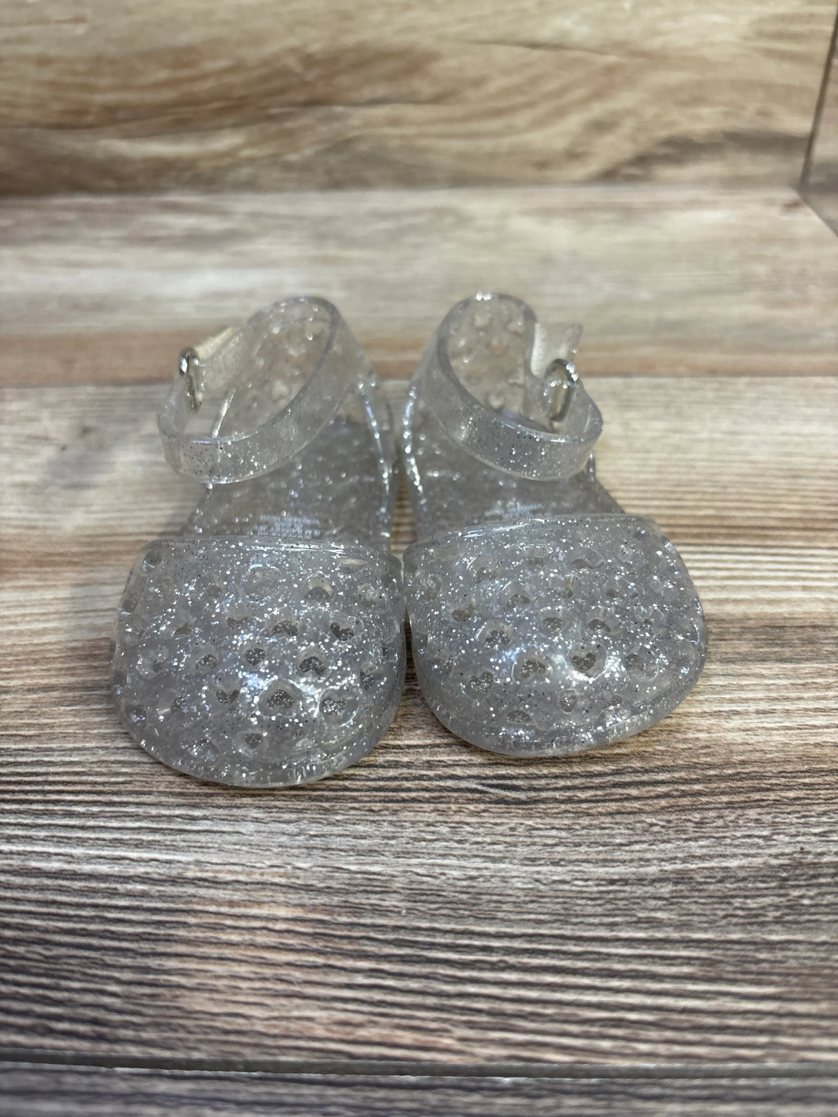 Front view of Old Navy clear glitter jelly toddler sandals showcasing heart-shaped toe holes and ankle strap on wood surface