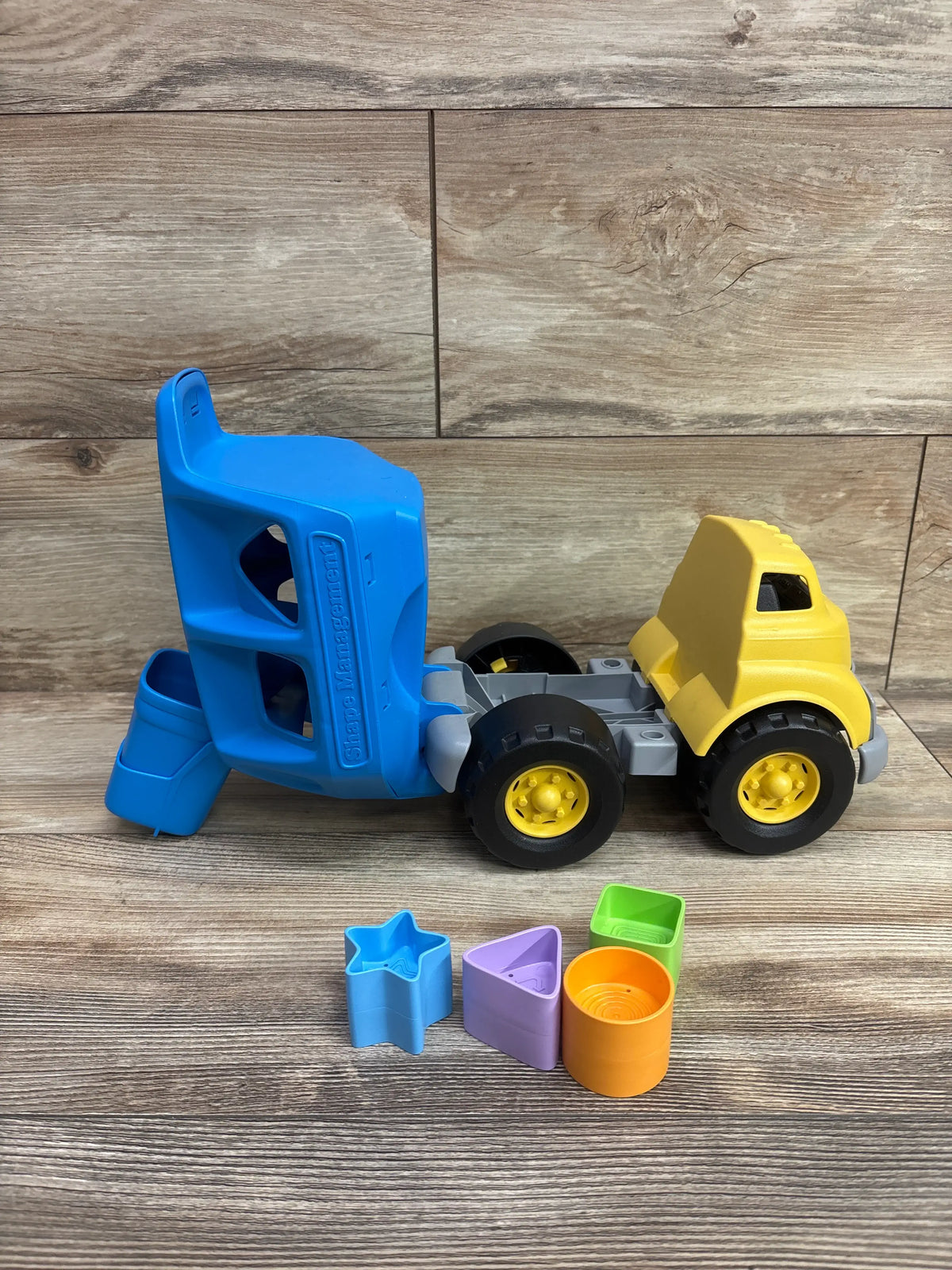 Side view of Green Toys Shape Sorter Truck with blue sorter body, yellow cab, and four colorful shape pieces on a wood background