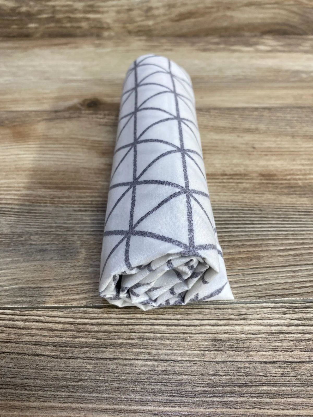 Front view of Graco Travel Lite crib sheet rolled to show white fabric with grey geometric grid pattern on wood surface