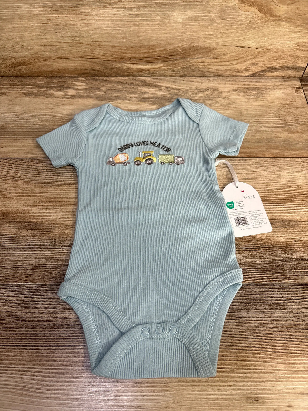Front view of the Kyle + Deena blue ribbed bodysuit showcasing its Daddy Loves Me A Ton truck design on a wood background