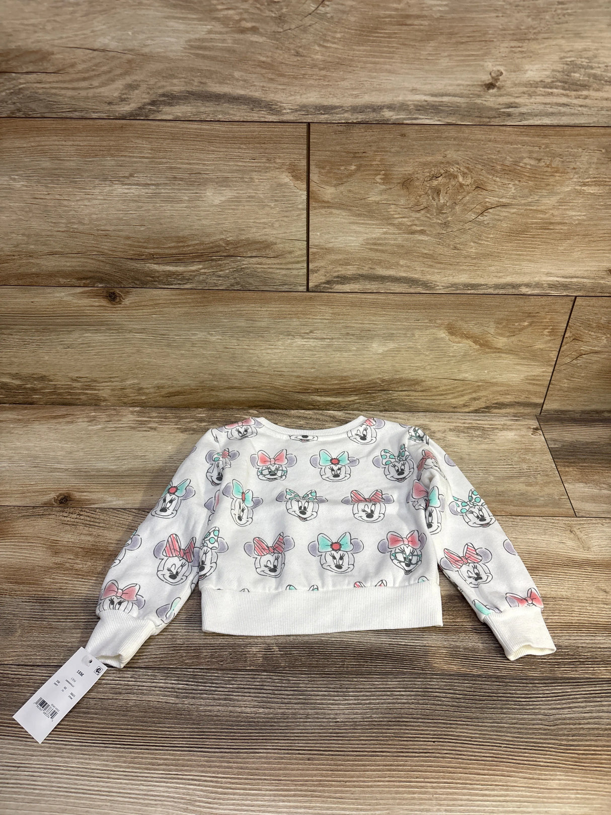 Flat-lay back view of white Disney Baby Minnie Mouse pullover sweatshirt showing all-over Minnie faces with pastel bows, ribbed hem, size 18M on wood background