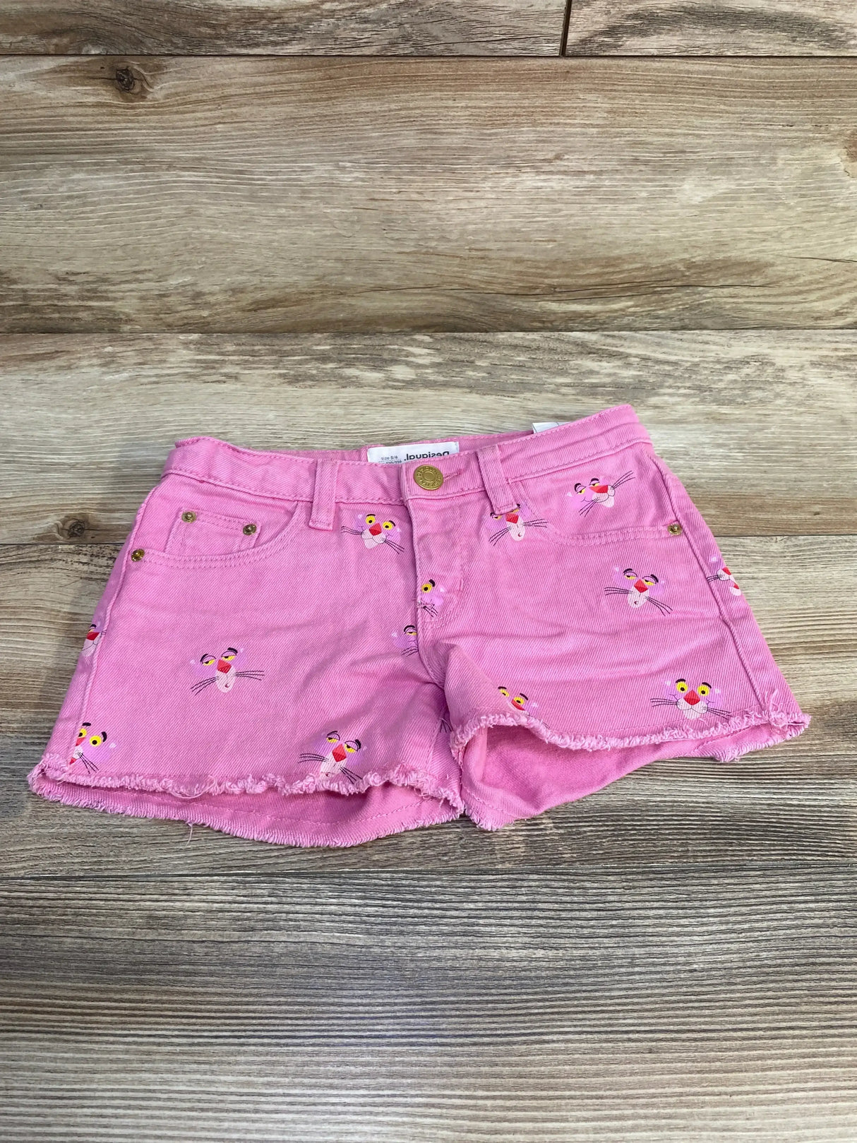 Front view of pink Desigual toddler shorts with Pink Panther face embroidery, frayed hem and gold button on wood background