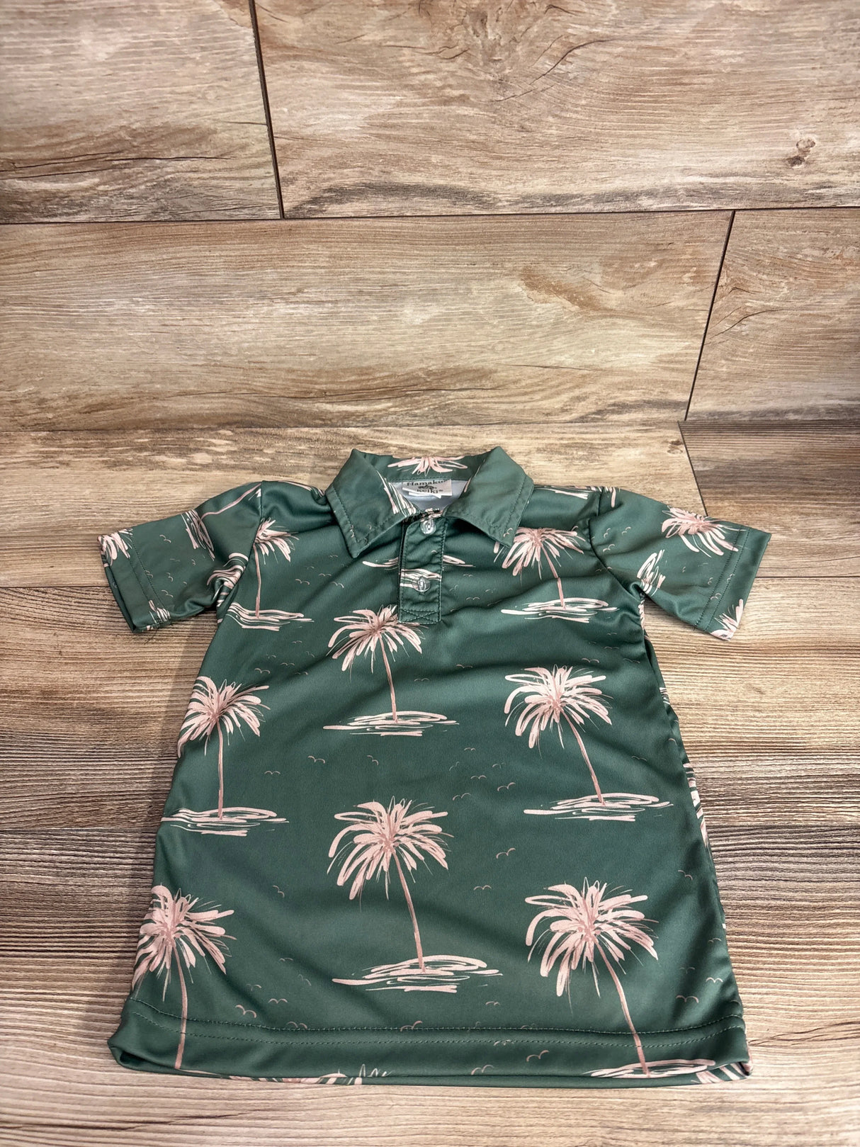 Flat lay of green Hamakua Keiki palm-print toddler polo shirt (size 3T) showing short sleeves and two-button placket on wood background