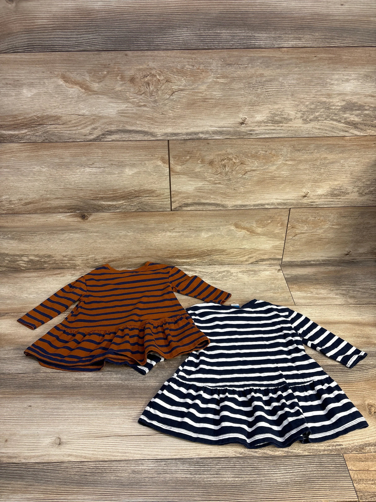 Two long sleeve striped toddler dresses, one brown with navy stripes, one navy with white stripes, on wood floor.