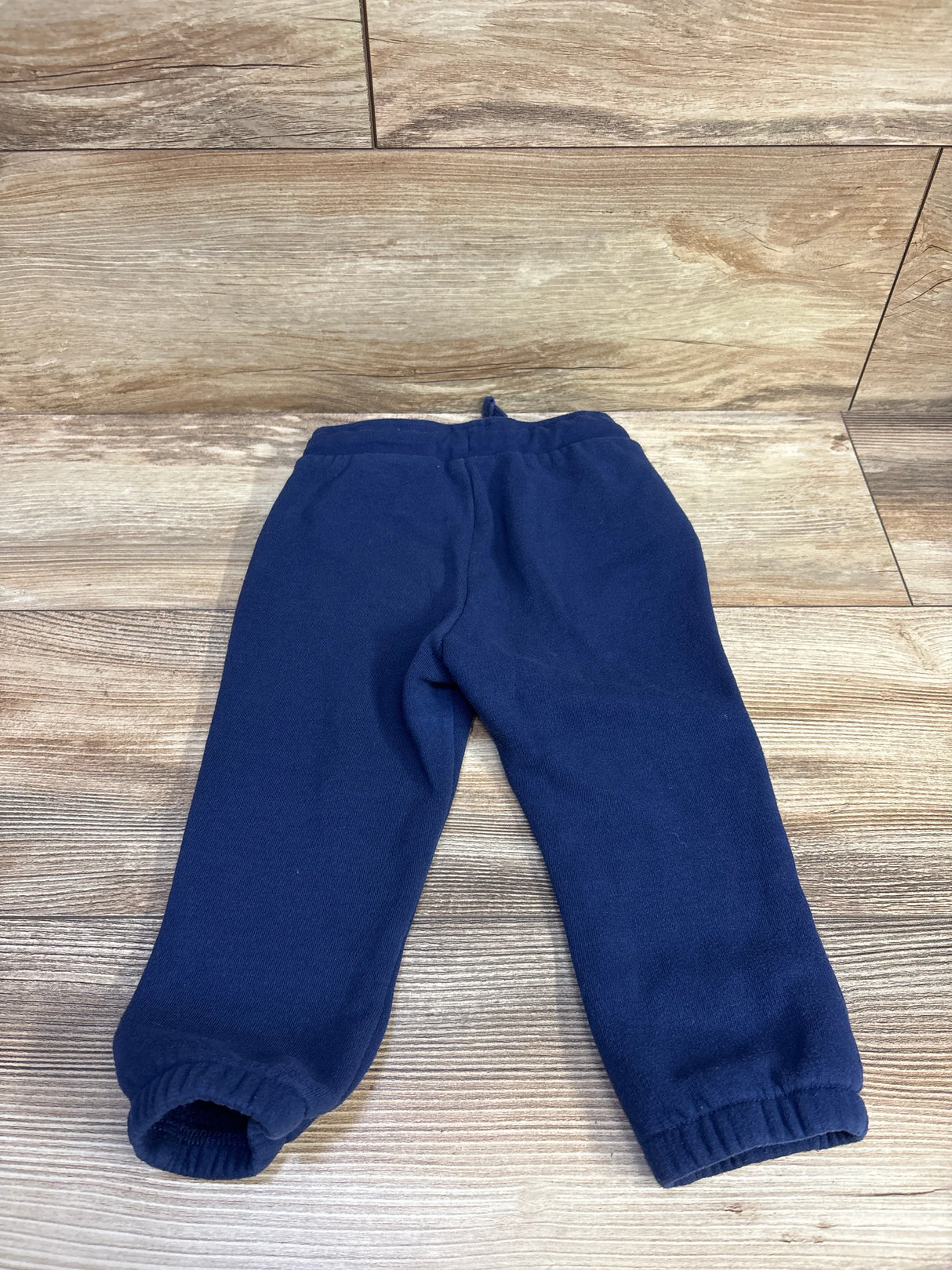 Laid-flat view of Okie Dokie navy toddler drawstring joggers showcasing elastic cuffs and drawstring waist on wood plank background, 18 months