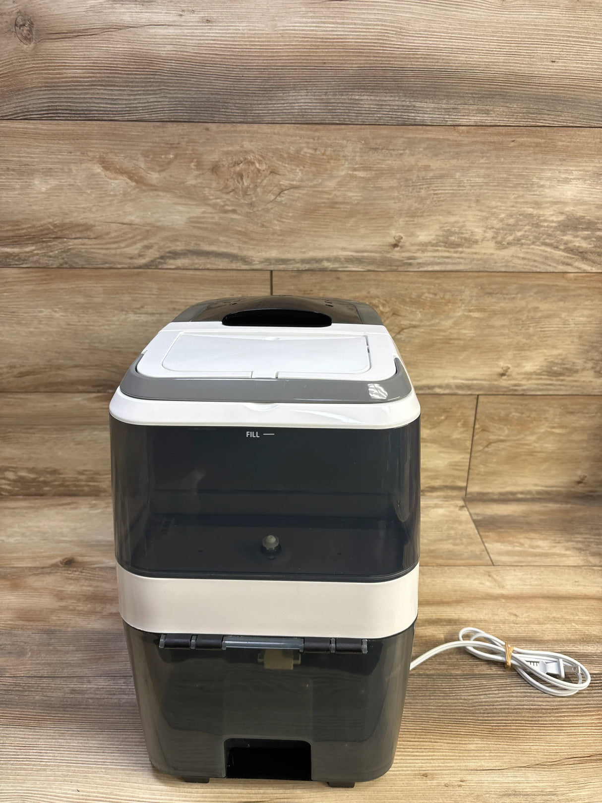 Front view of Baby Brezza Bottle Washer Pro showing transparent water tank, top lid, and coiled power cord on wood backdrop