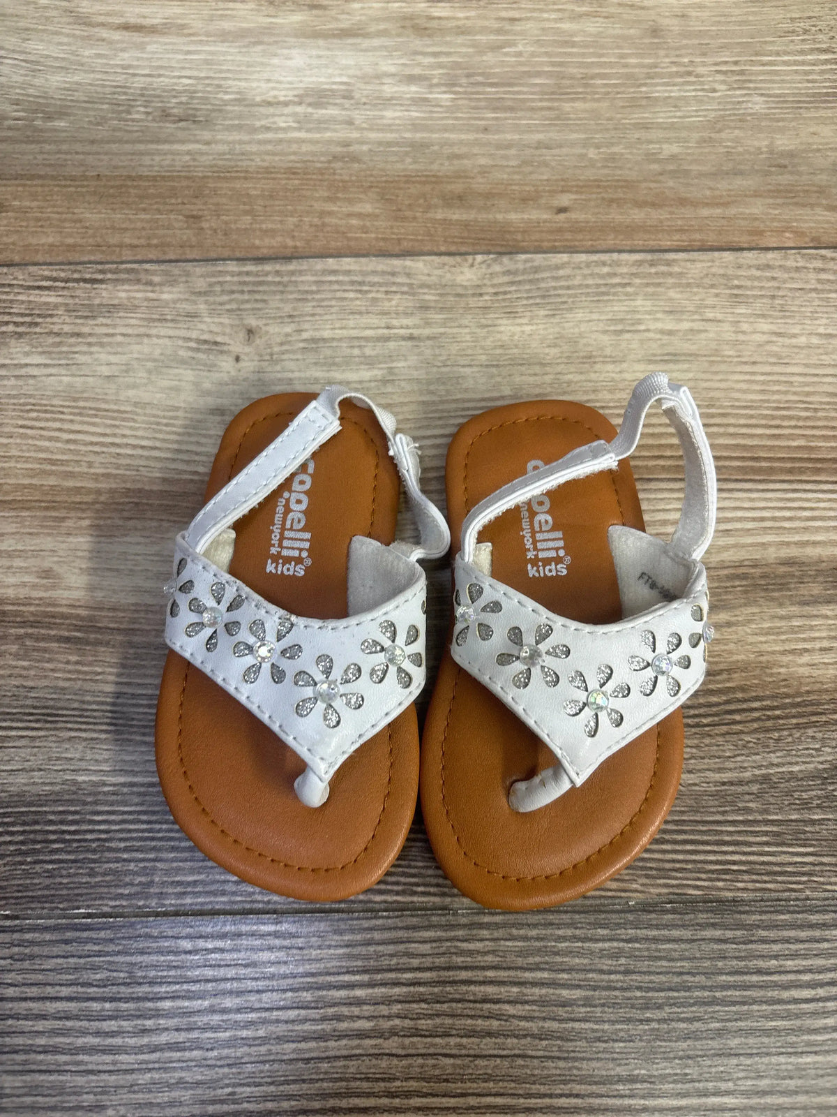 Top view of white Capelli backstrap sandals, size 4.5C, showcasing silver flower embellishments on a wood surface