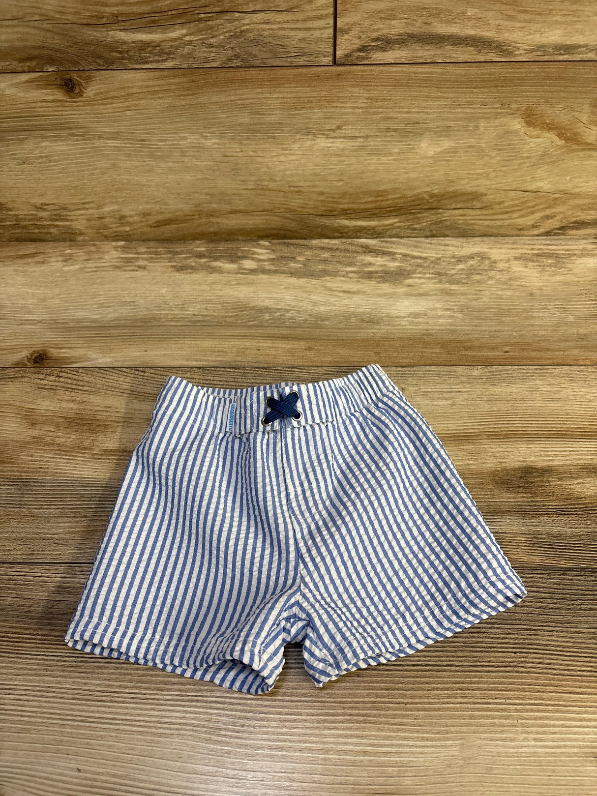 Flat lay of Rugged Butts seersucker striped toddler swim shorts showing elastic waistband with navy drawstring on wood background