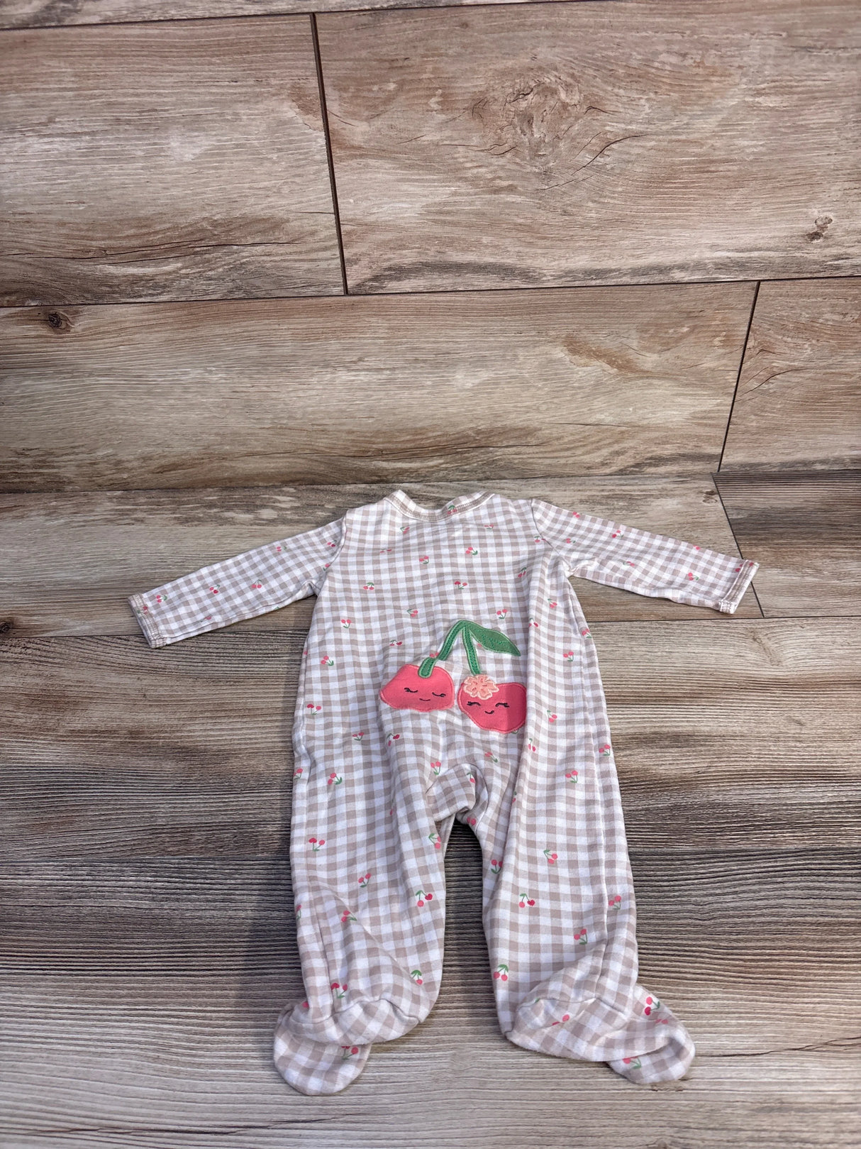 Flat lay of beige gingham Carter's footed baby sleeper size 3M showing cherry appliqué on front with wood floor background