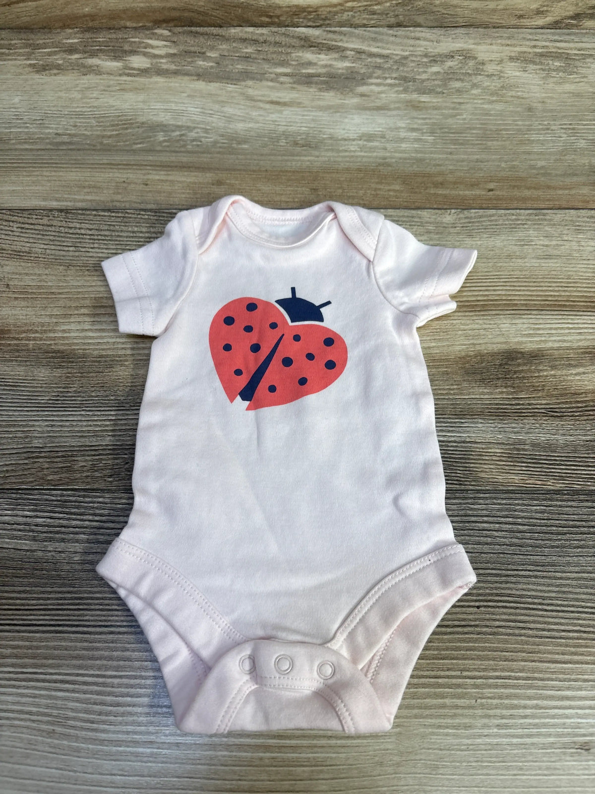 Front view of Primary pink newborn ladybug bodysuit showcasing ladybug print and snap closures on wood-grain background