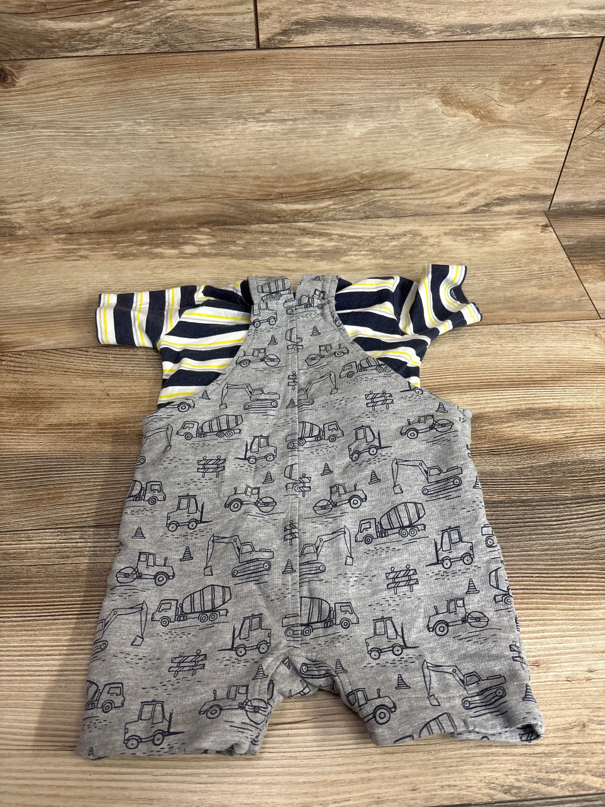 Flat lay of baby 2-piece: navy and yellow striped shirt with gray shortalls featuring construction vehicles on wood floor