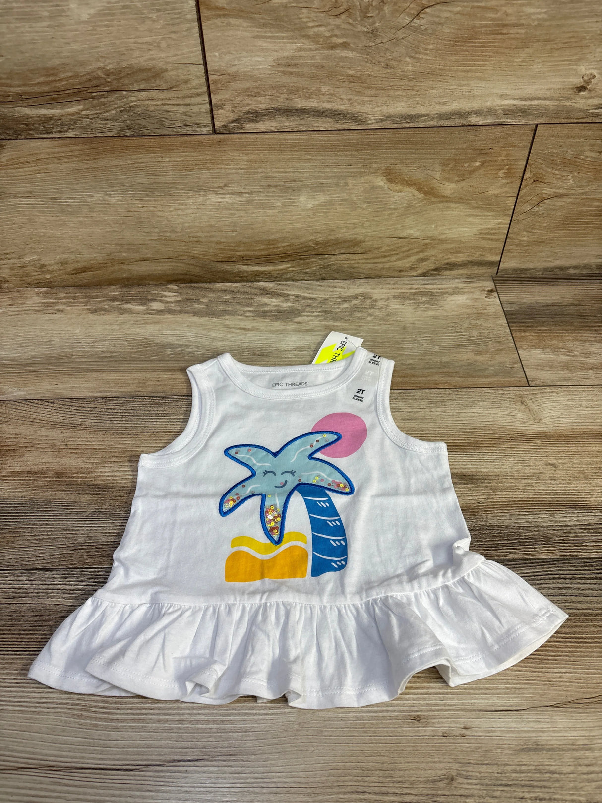 Flat lay of Epic Threads white peplum tank top (2T) showcasing smiling starfish graphic and ruffled hem on wood background