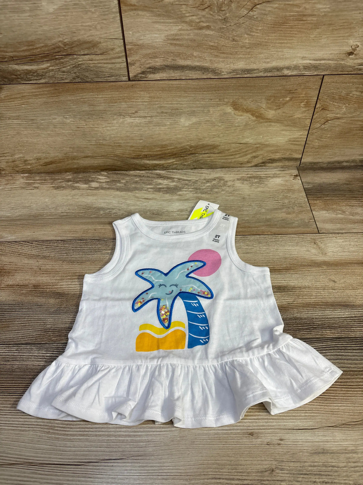 Flat lay of Epic Threads white peplum tank top (2T) showcasing smiling starfish graphic and ruffled hem on wood background