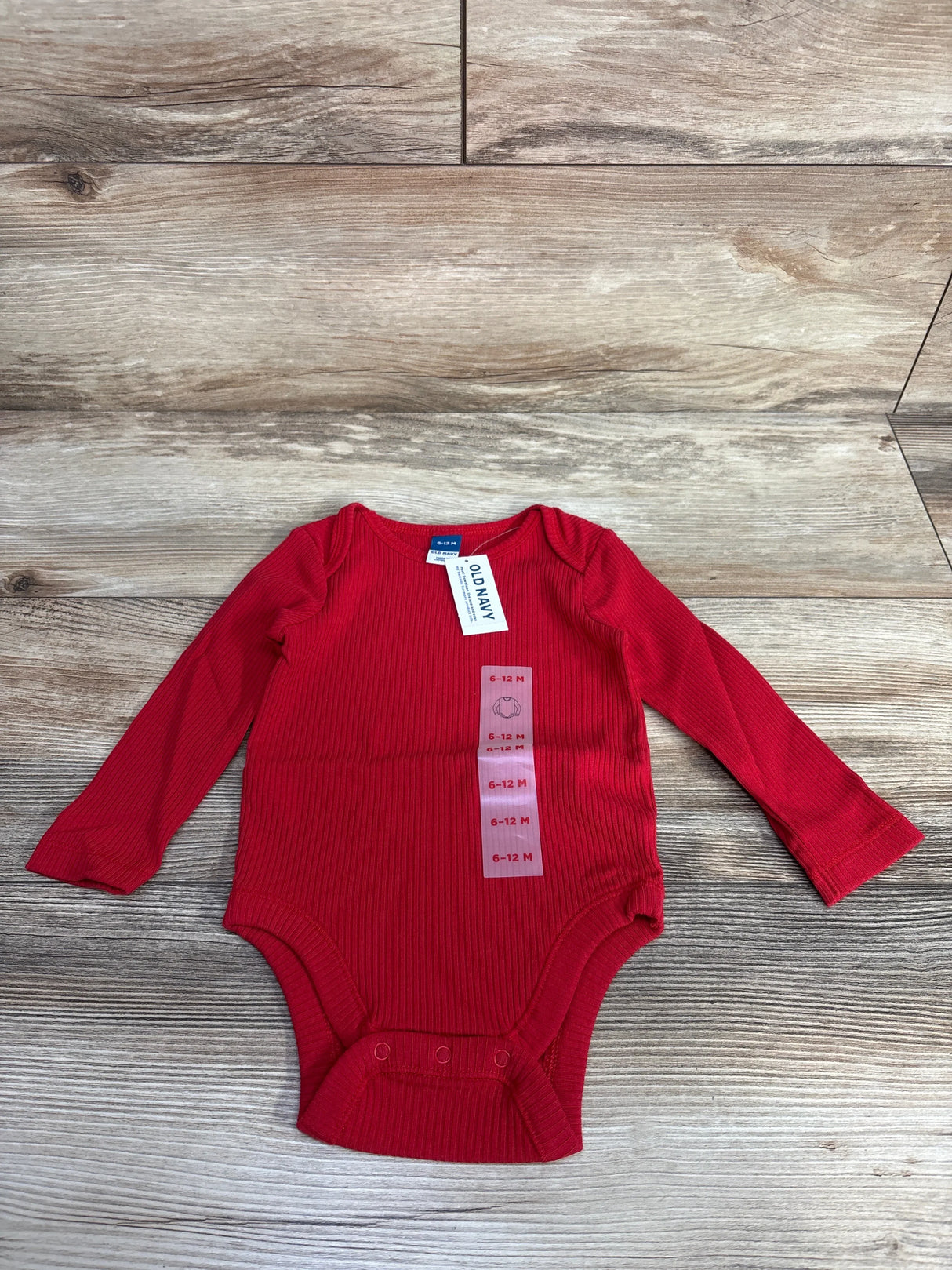 Flat lay of NWT Old Navy red ribbed long sleeve baby bodysuit size 6-12m showcasing hangtags and snap closure on wood plank floor
