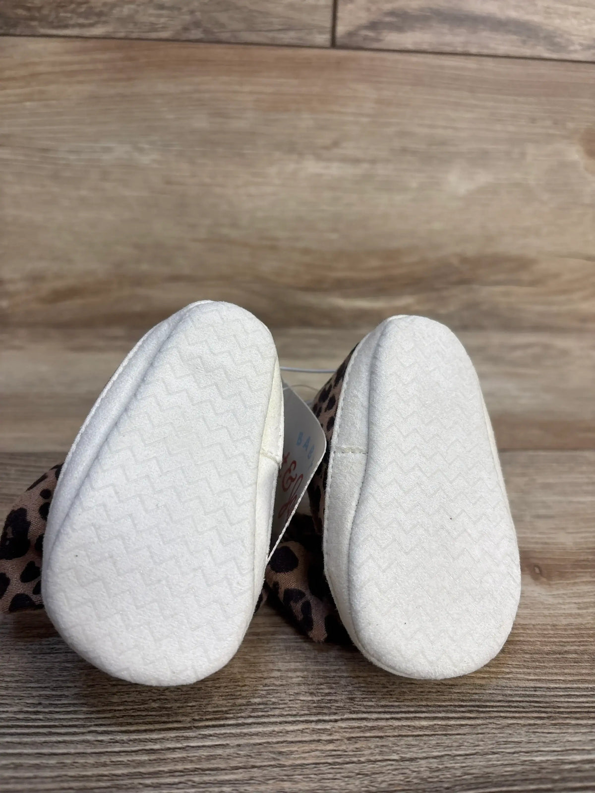 Bottom view of Cat and Jack leopard bow crib sneakers showing soft white soles and leopard-print fabric bows on wood backdrop