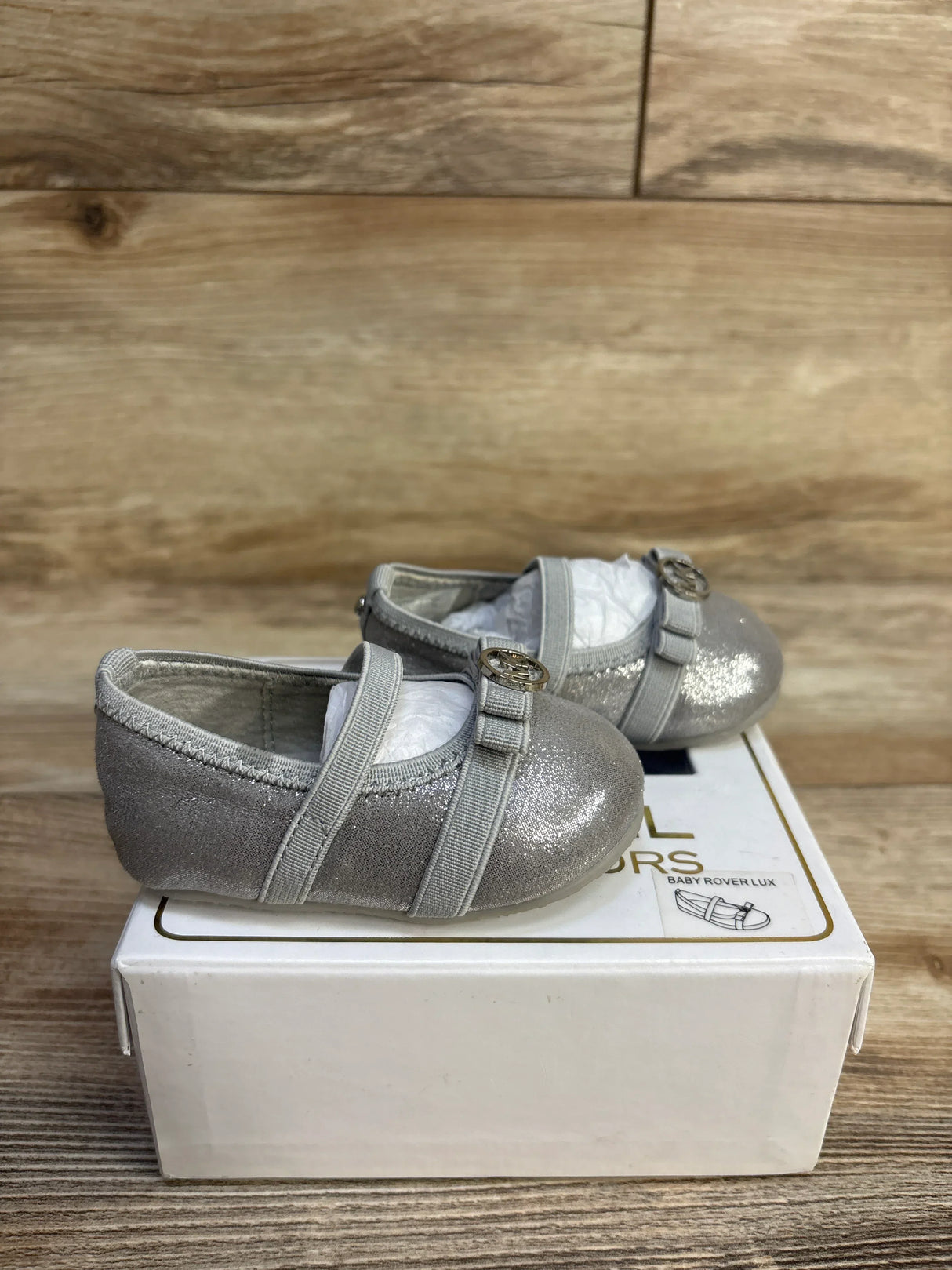 Side view of Michael Kors Baby Rover Lux silver baby shoes (size 2C) showcasing glitter finish and MK logo strap, resting on white shoebox on a wooden surface