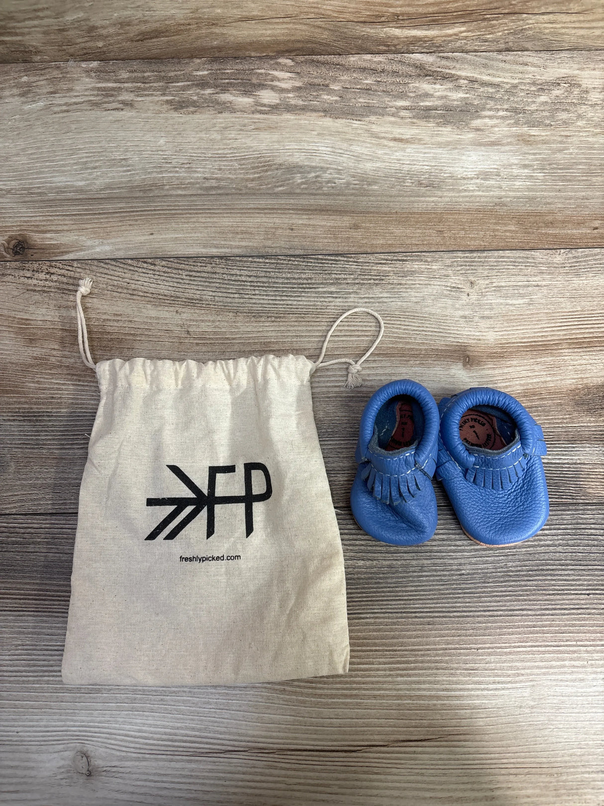 Blue leather baby moccasins and Freshly Picked drawstring bag on wood background