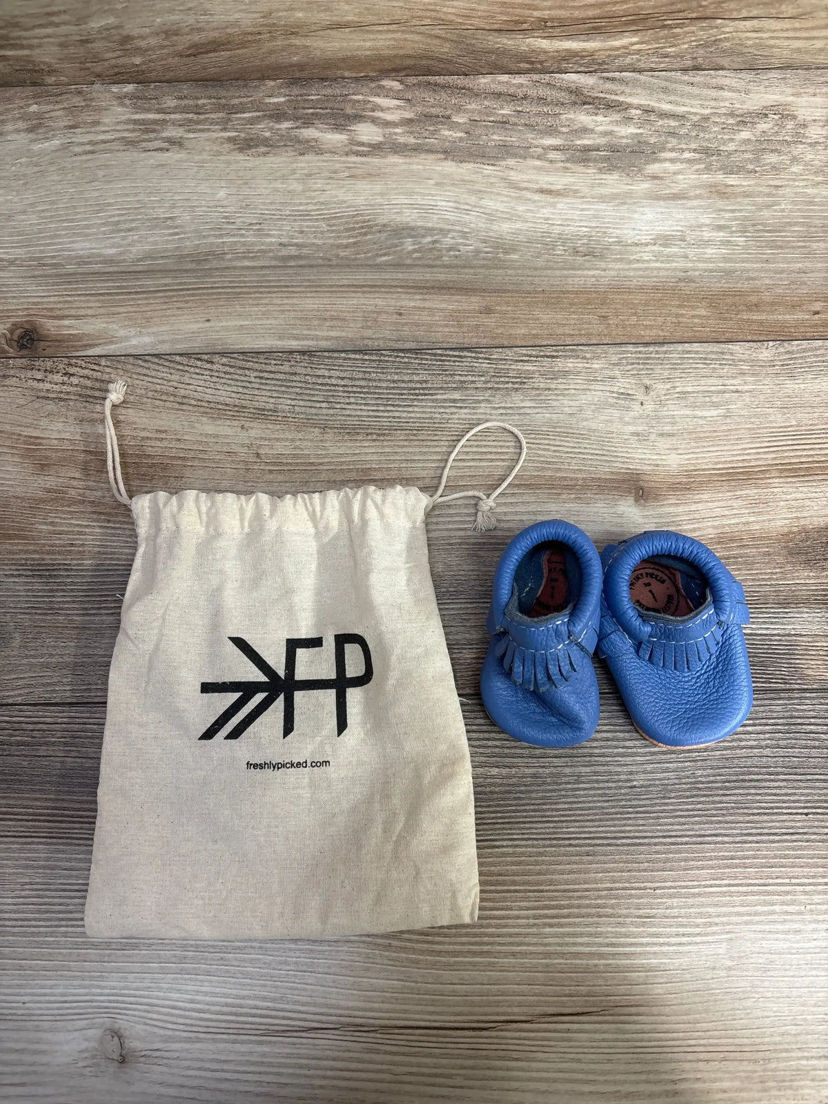 Blue leather baby moccasins and Freshly Picked drawstring bag on wood background