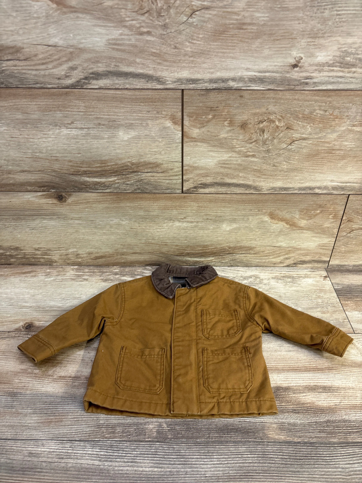 Front flat lay of baby tan canvas barn coat with brown corduroy collar and front patch pockets on wood-plank background