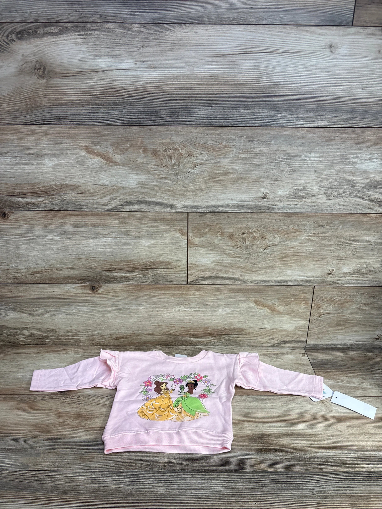 Front view of pink baby sweatshirt (3-6m) showcasing Belle and Tiana graphic and ruffled shoulders on wood floor