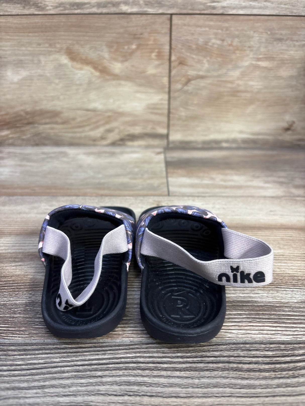 Rear view of Nike Kawa Slide SE TD sandals showing butterfly-print pink strap and heel strap with Nike logo on wood floor