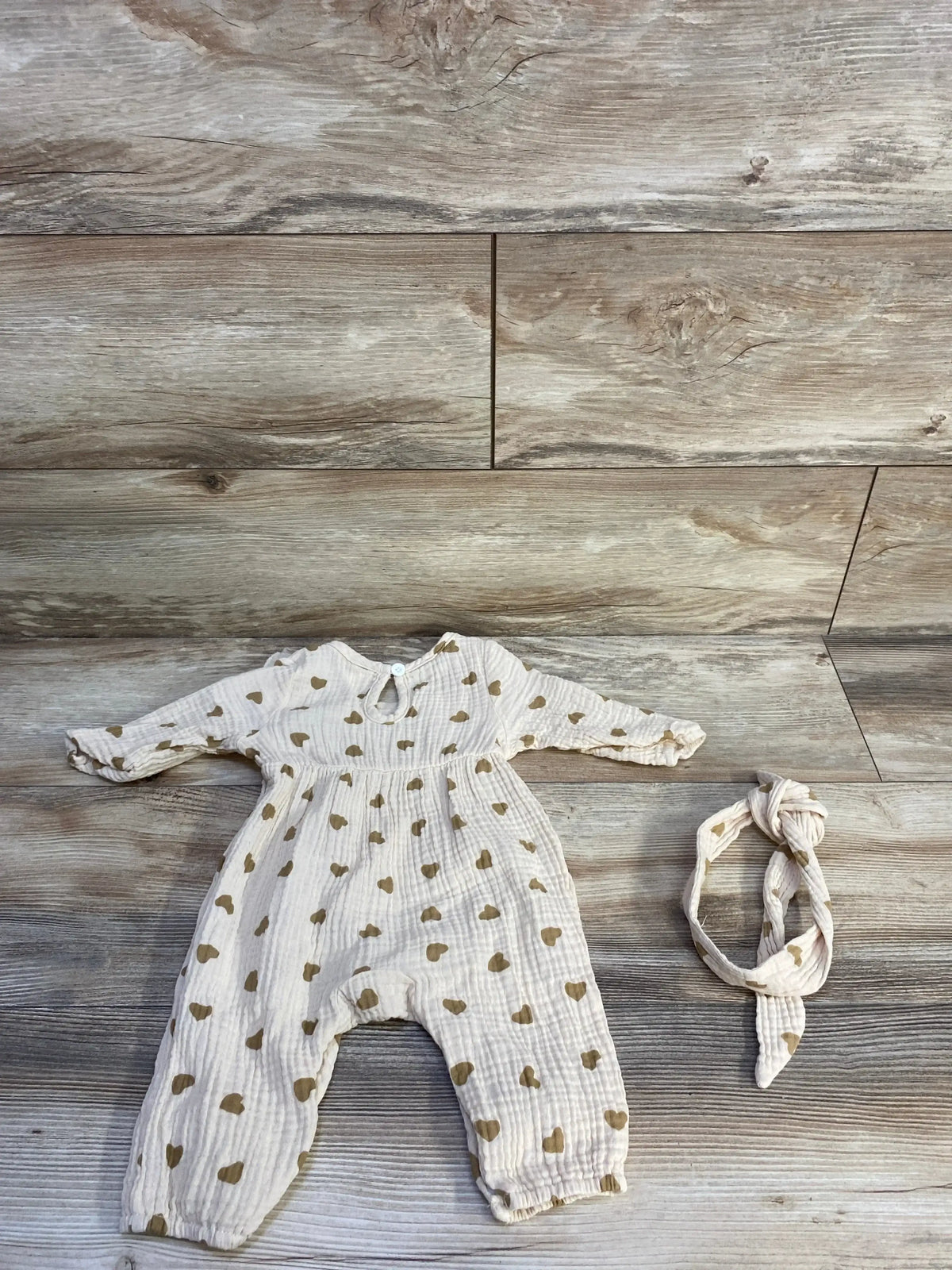Flat lay of beige gauze baby jumpsuit and matching headband showing gold heart print, long sleeves and gathered cuffs on wood background
