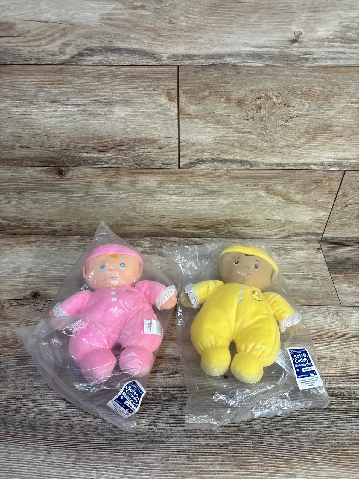 Flat lay of NEW Lakeshore Cuddly Washable Dolls in pink and yellow sleeper outfits in sealed packaging on wood plank floor