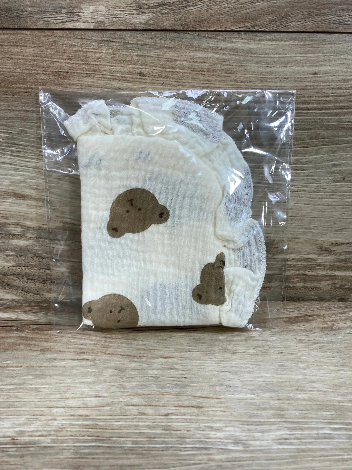 Folded front view of cream ruffle cotton baby bib with brown bear print in clear plastic packaging on wooden surface
