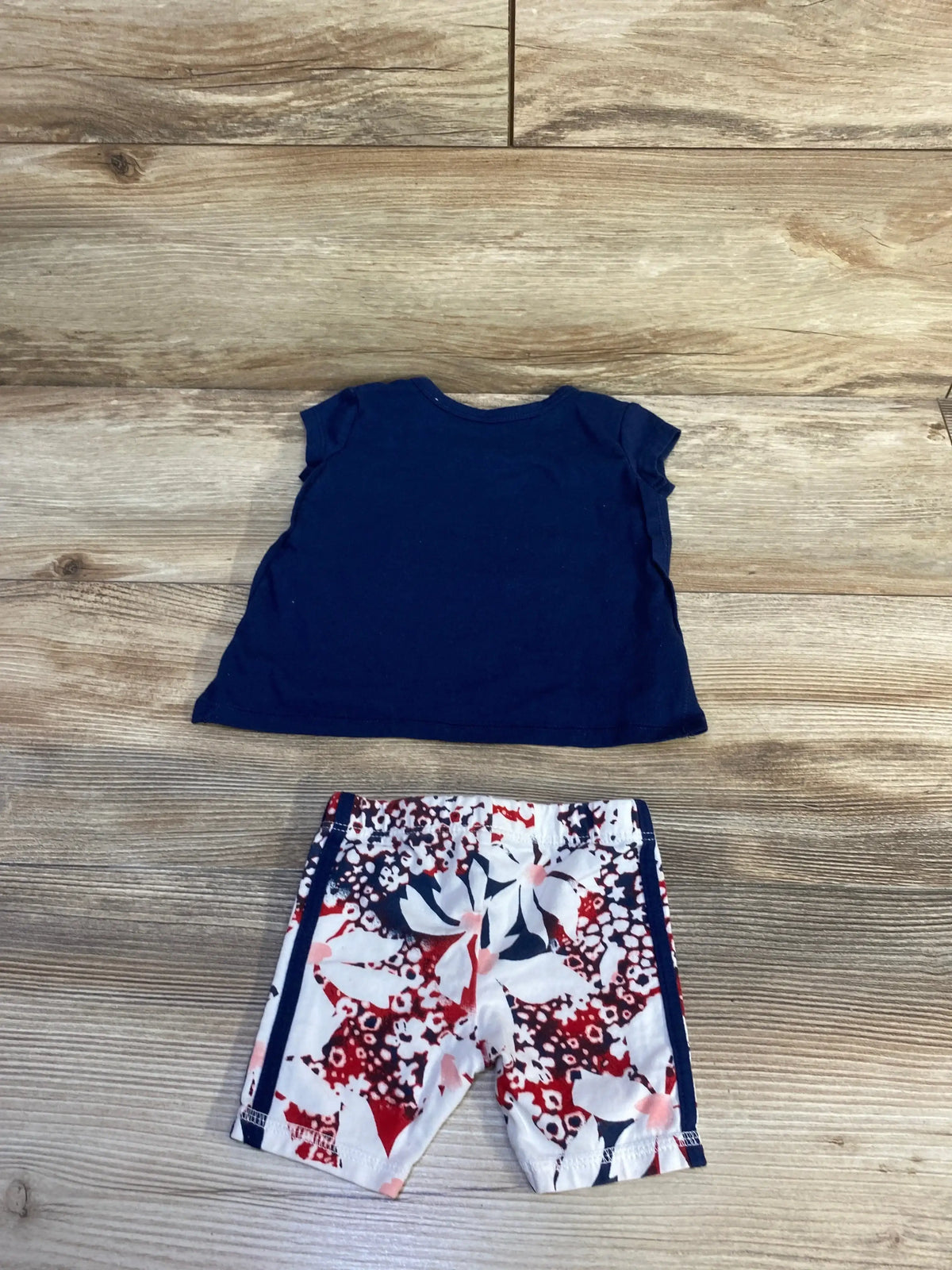 Flat lay of Adidas two piece floral logo shirt and shorts navy size 3m showcasing solid navy tee and red white blue floral bike shorts on wood plank floor