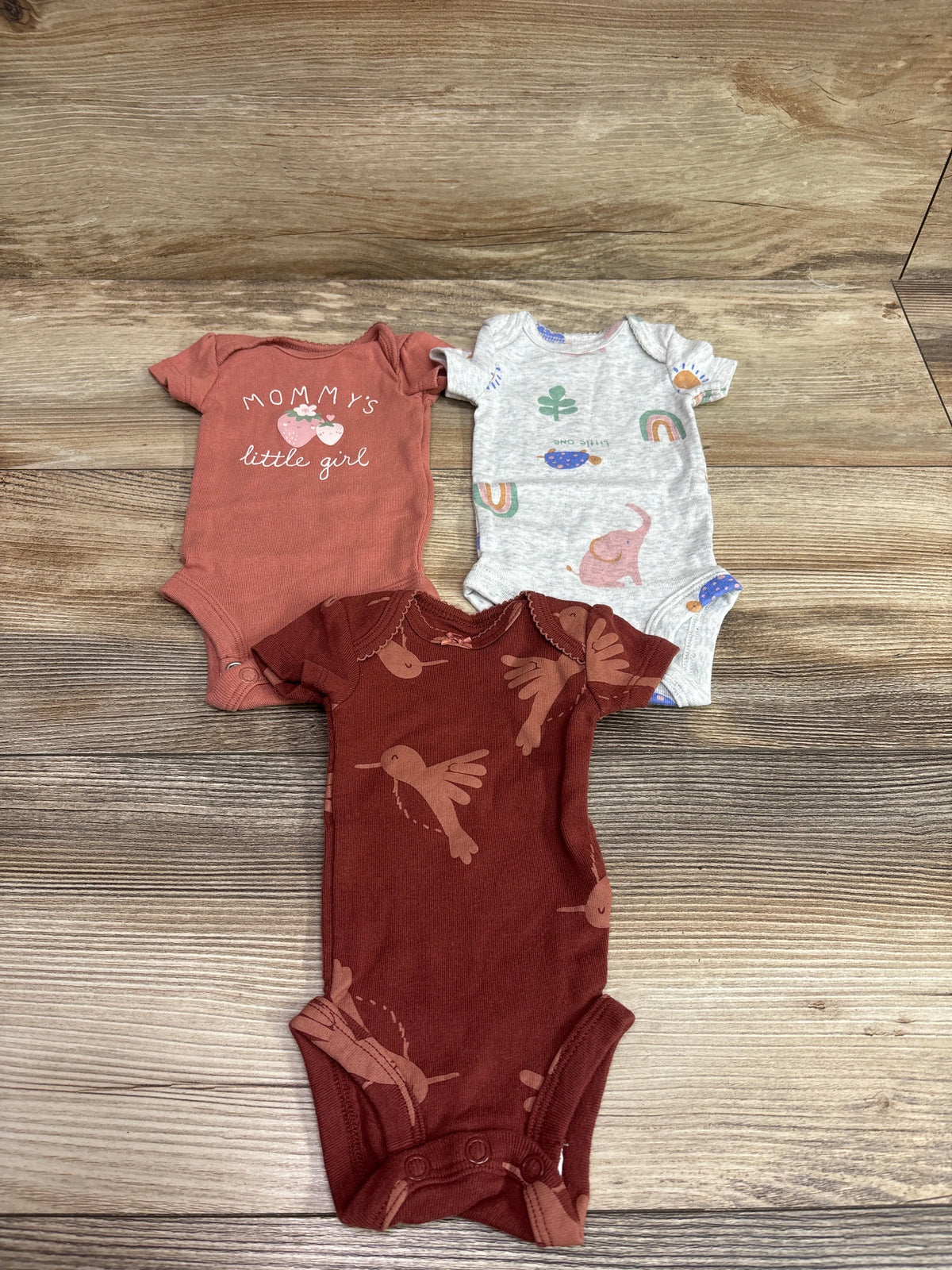Top-down view of Carter’s 3-pack preemie bodysuits in pink, gray, and brown with printed designs on a wood surface