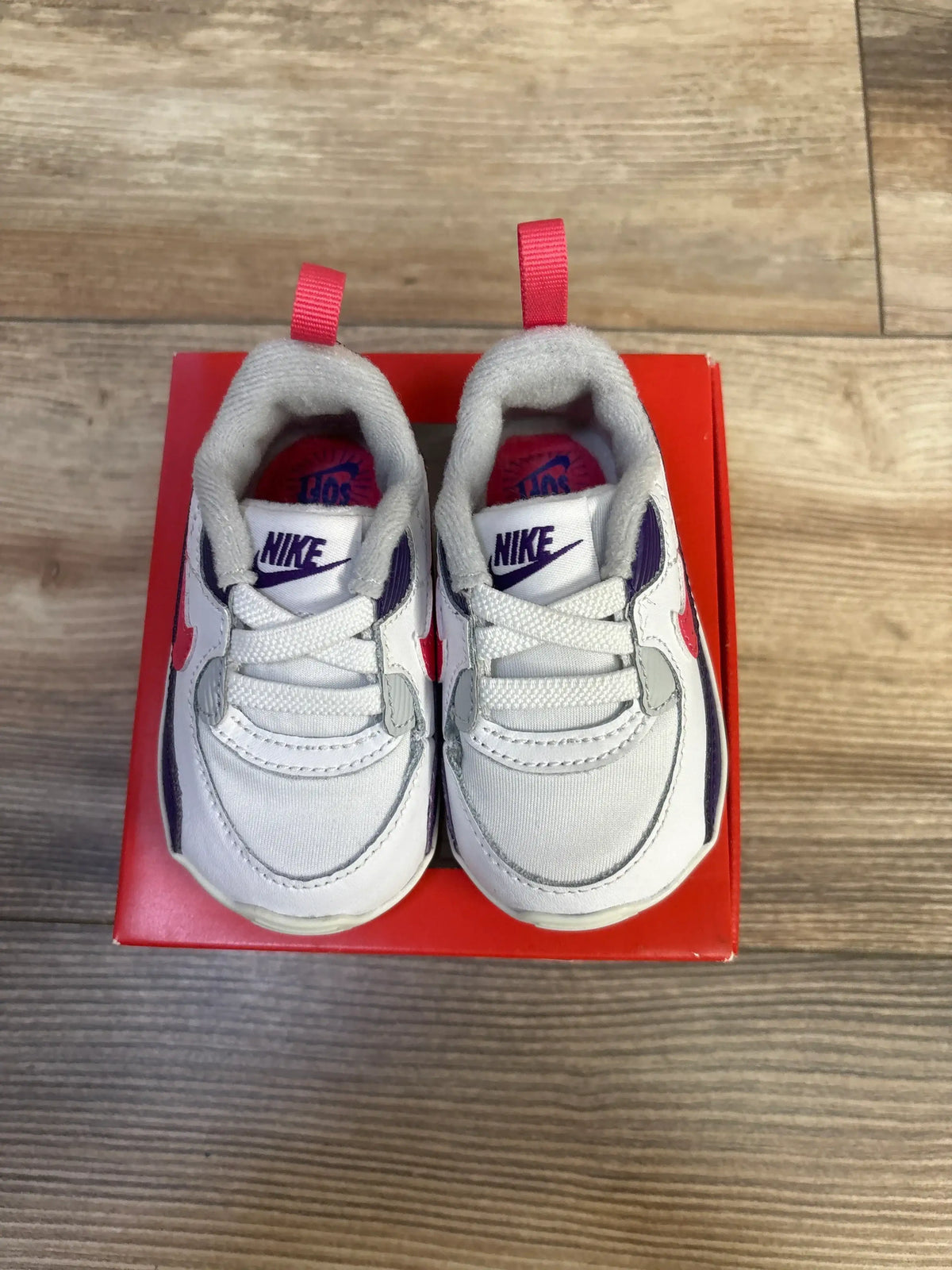 Overhead view of Nike Air Max 90 crib sneakers, white with pink accents and grey overlays, purple tongue logos, on red box