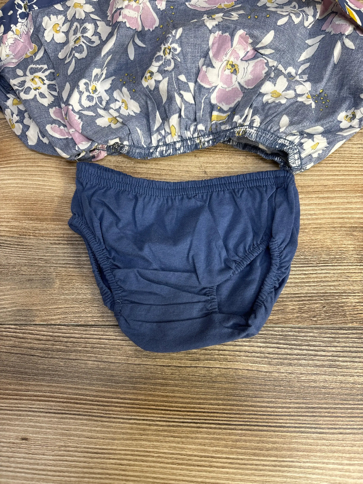 Flat lay of Just One You blue floral baby dress with navy bloomers showing elastic waistbands on wood background