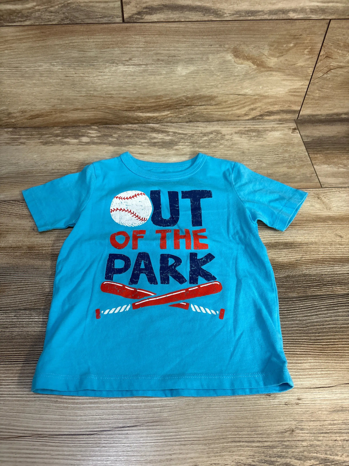 Front view of Children’s Place blue toddler shirt, size 3T, showcasing an OUT OF THE PARK baseball graphic on a wood floor