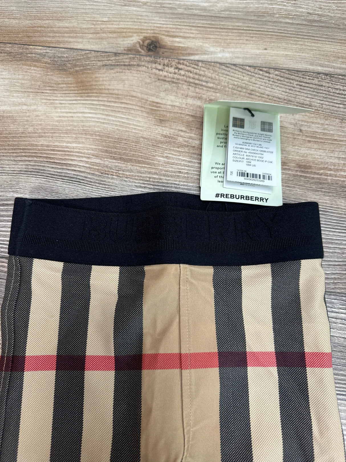 Top view of Burberry 18M baby leggings showing beige check pattern, black elastic waistband embossed Burberry, and tag on wood floor