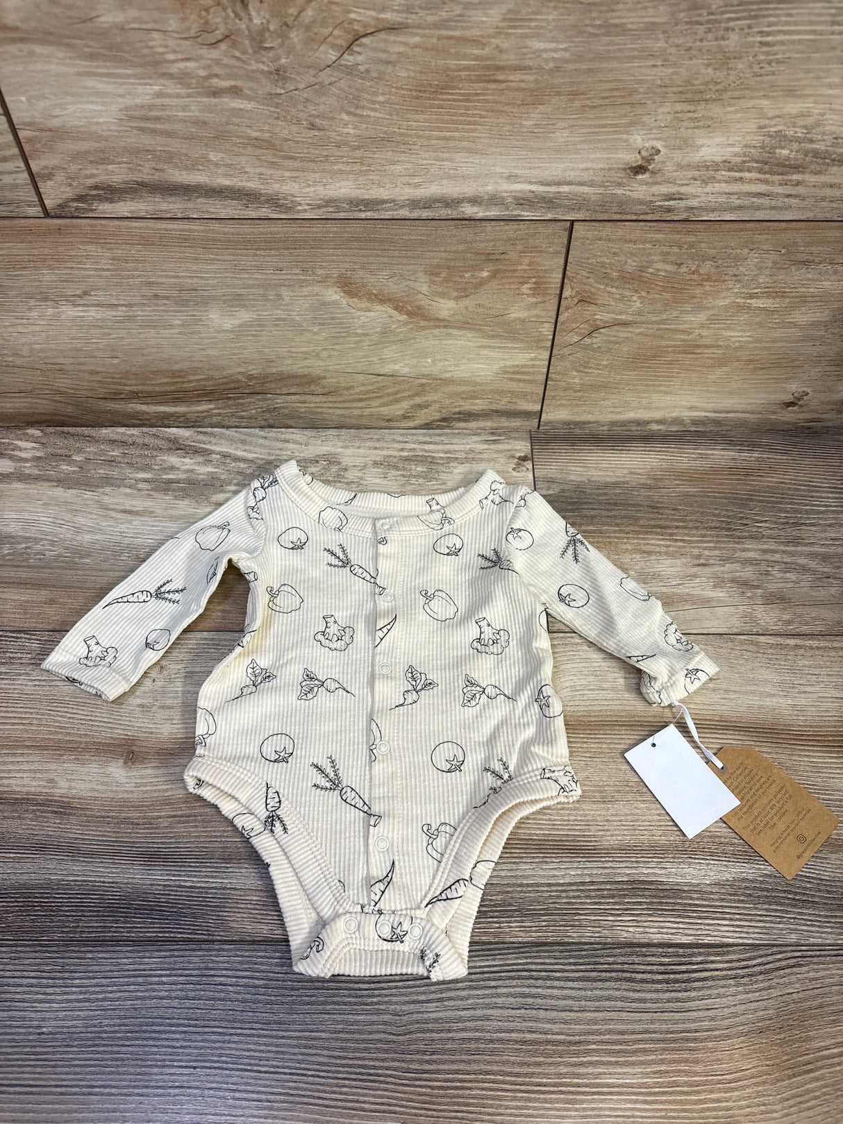 Flat lay of ivory ribbed long-sleeve baby bodysuit showcasing front snap closures and vegetable line-print on wood background, 0-3M