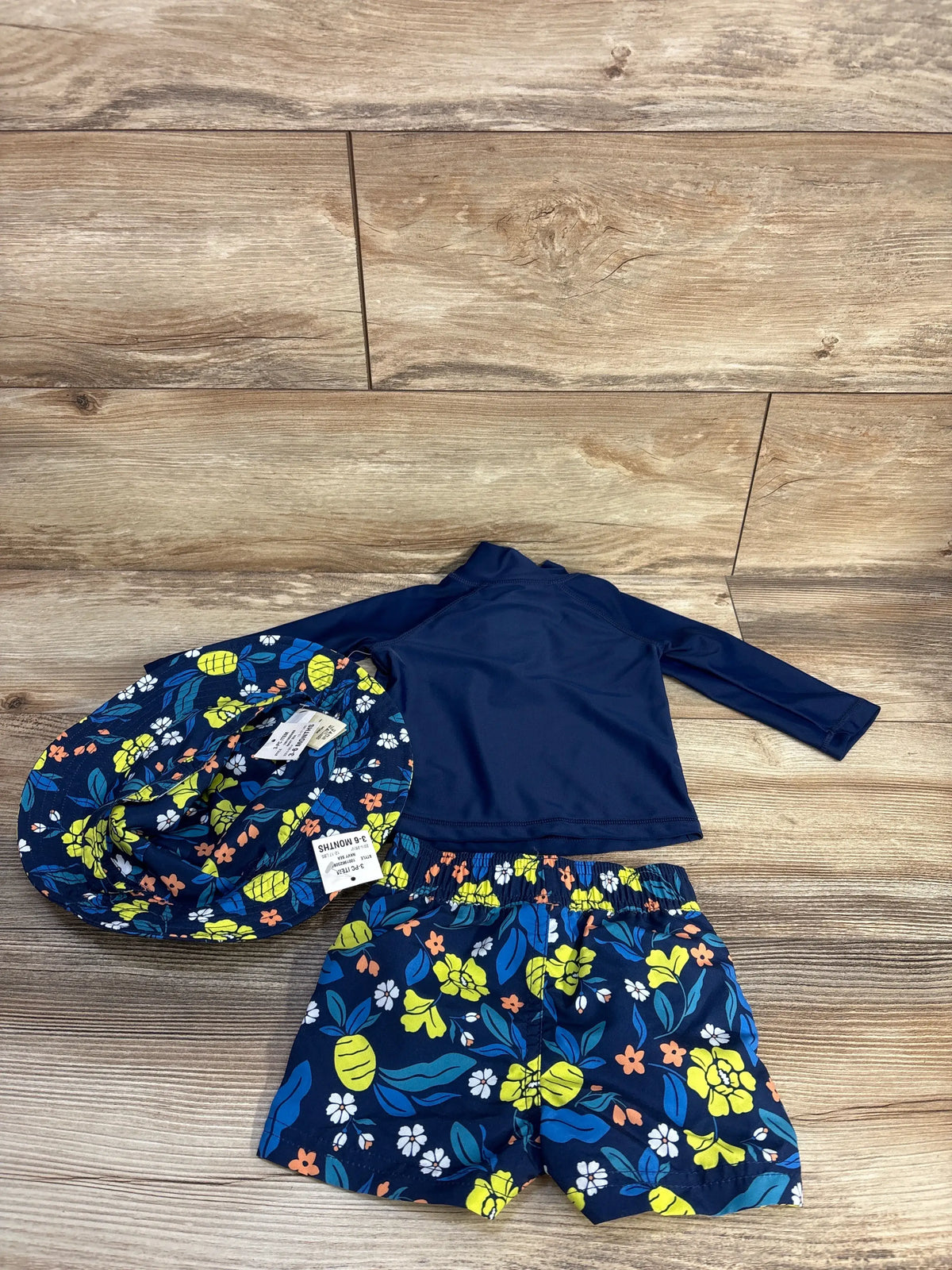 Flat lay of First Impression navy three-piece swim set with floral trunks, long-sleeve rash guard, and matching hat