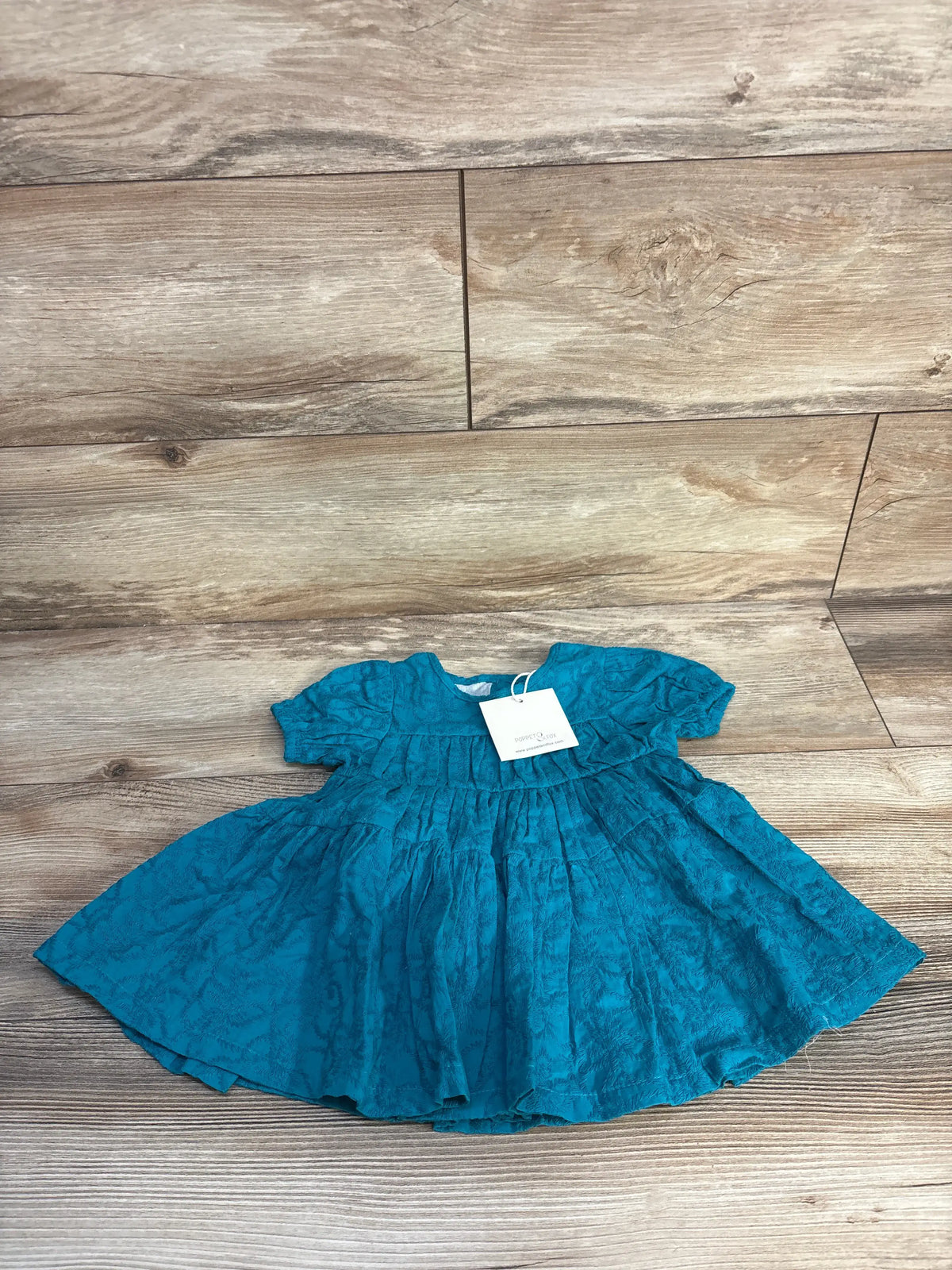 Front view of Poppet & Fox Sweet Party Dress showcasing blue textured fabric, gathered skirt, and short sleeves on a wood floor
