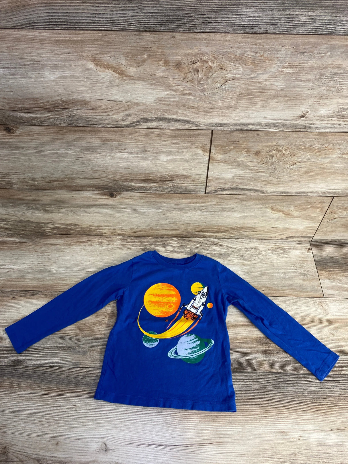 Blue long sleeve kids shirt with colorful space rocket and planets graphic on wood floor