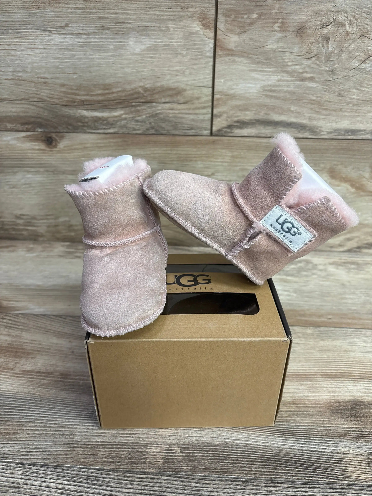 Pair of pink suede baby UGG boots with soft lining, displayed on a cardboard UGG box on wooden surface