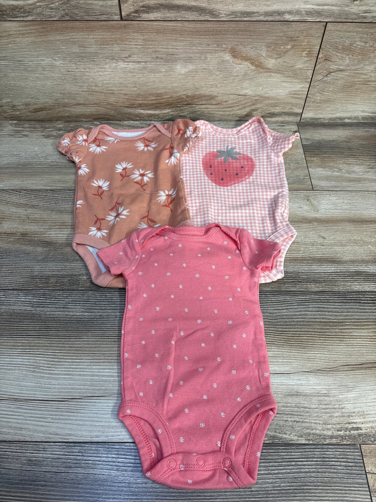 Top-down view of Just One You 3pk pink bodysuit set showcasing floral, strawberry, and daisy prints on wood flooring