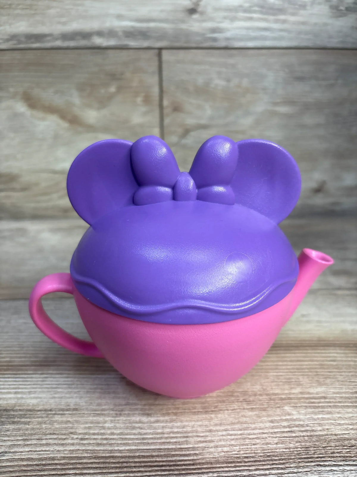 Front view of Green Toys Minnie Mouse teapot showing purple Minnie-ear lid with bow and pink teapot base on wood background