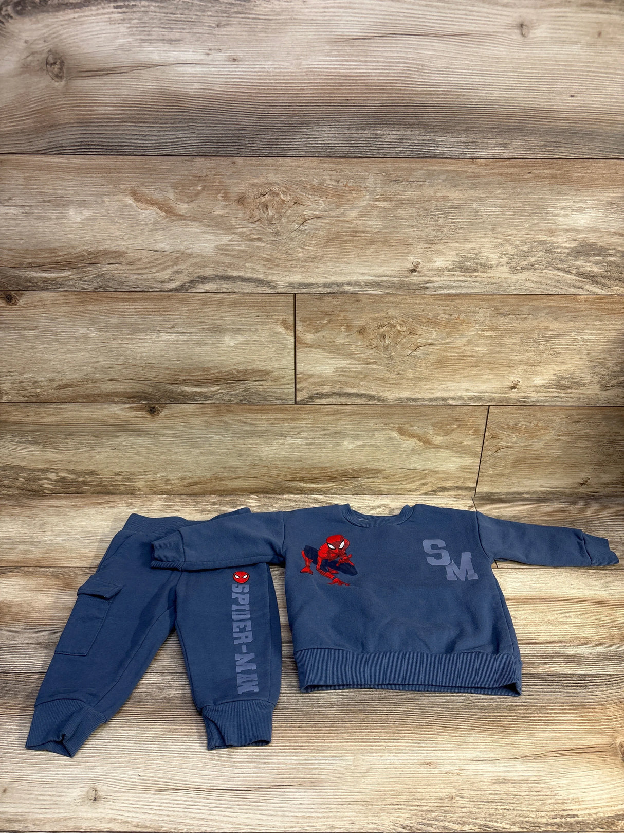 Front view of toddler blue Marvel Spider-Man sweatshirt and joggers showing red Spider-Man graphic and ‘SPIDER-MAN’ on pant leg on wood background
