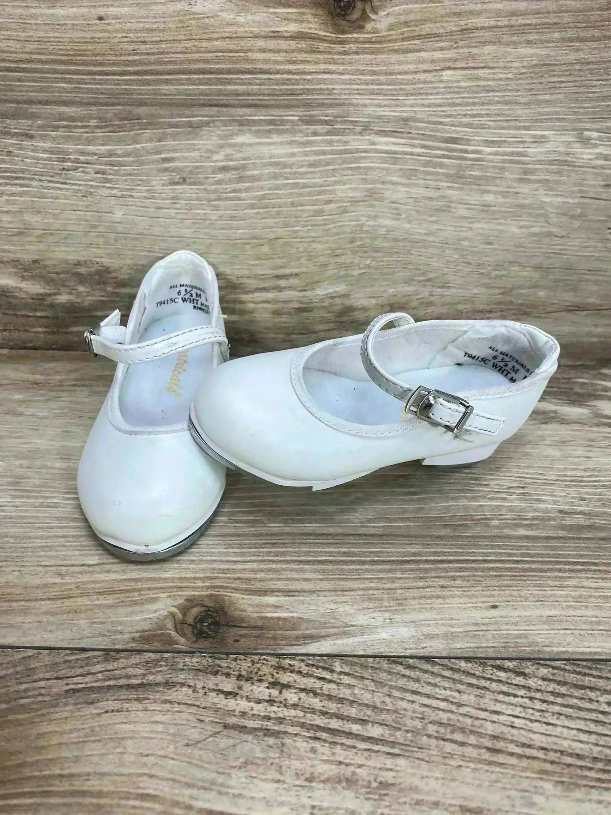 Angled front view of white Theatricals tap buckle toddler shoes showcasing buckle strap and metal taps on a wood plank background